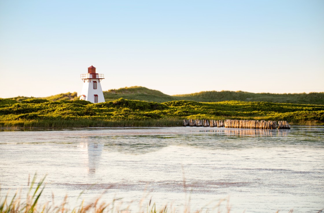 A white octagonal lighthouse with a red top stands on a grassy cliff overlooking a beach and calm sea.