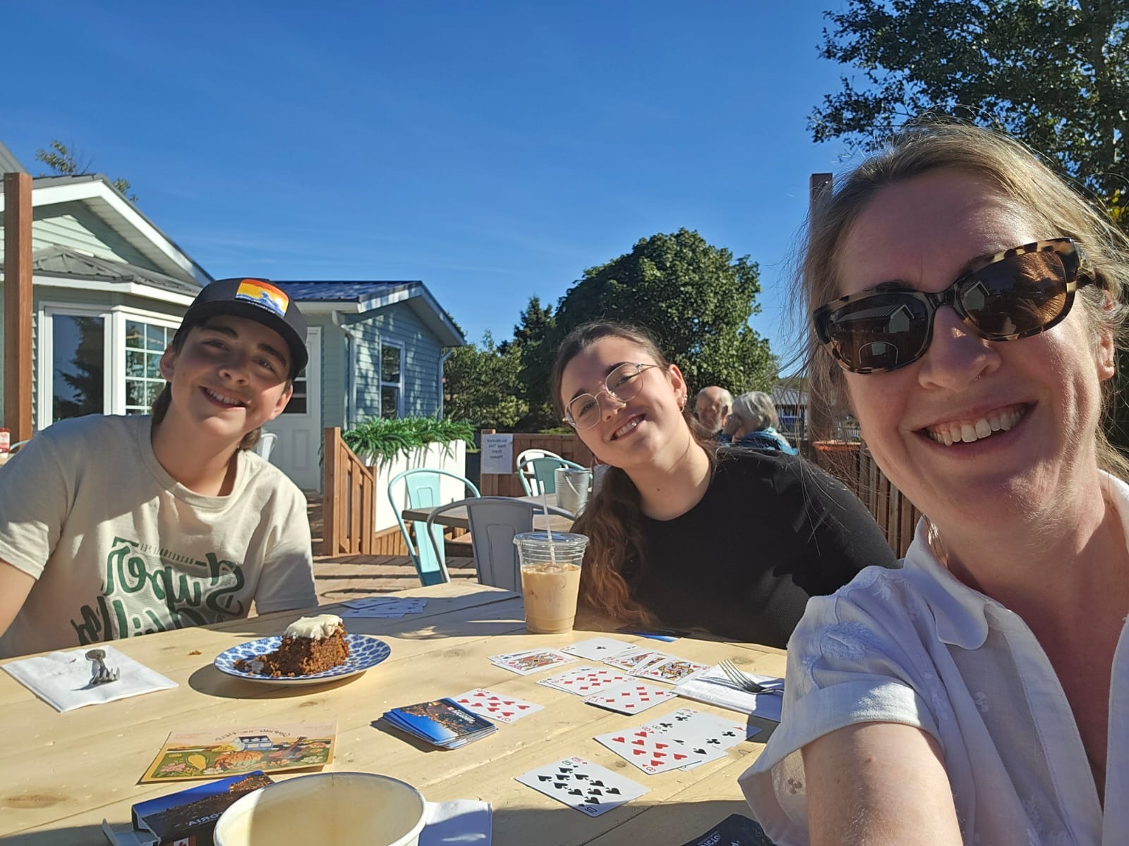 A group of people sit at an outdoor café table on a sunny day, drinking iced coffee, eating cake, and playing cards.