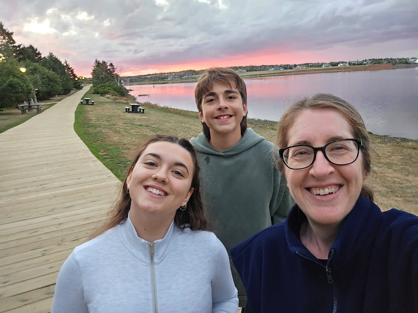 Three people taking a selfie on a boardwalk beside calm water at sunset