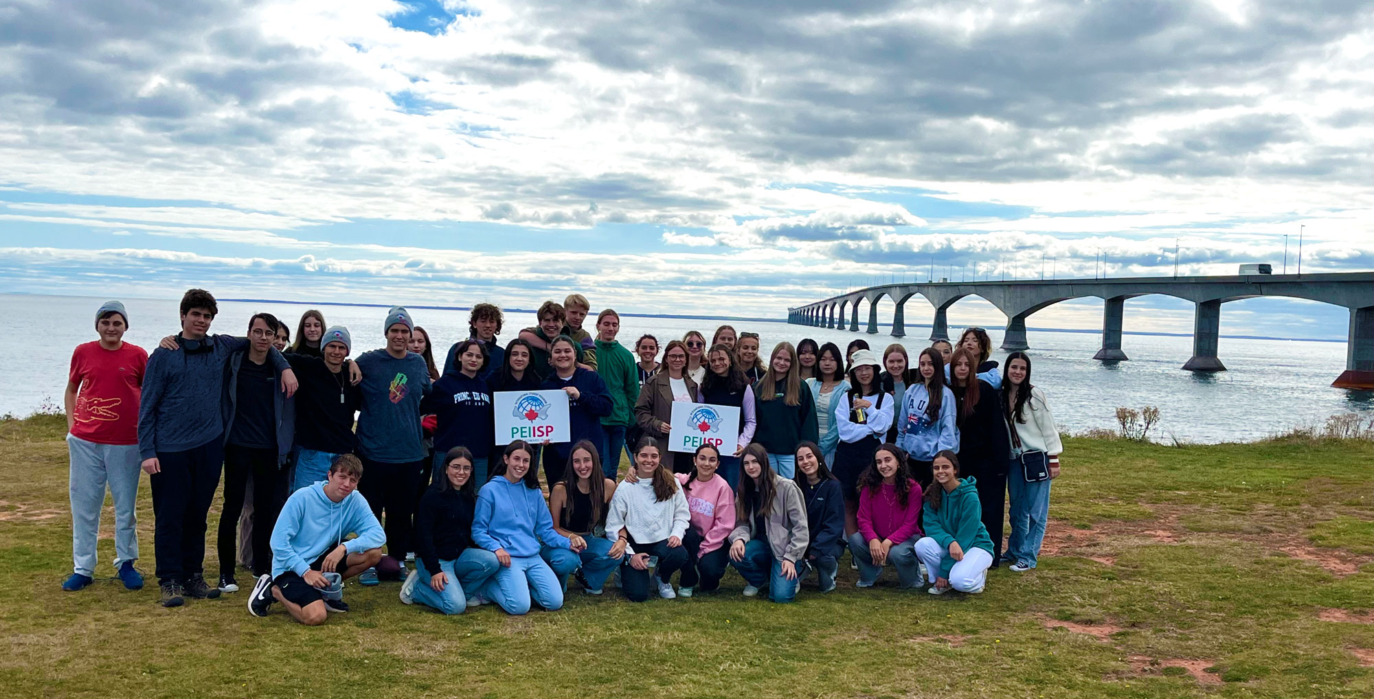 A large group of students poses together on a grassy shoreline with the Confederation Bridge stretching across the water in the background.